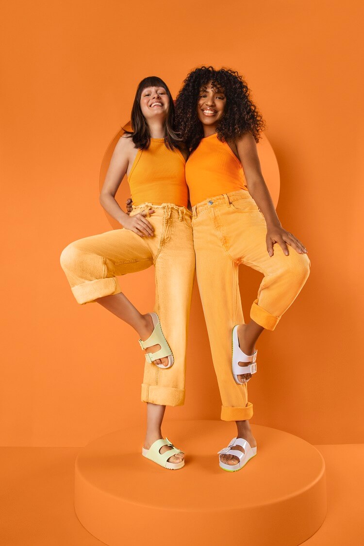 Duas mulheres em fundo laranja, usando roupas laranja e sandálias da coleção Zaxy + Gabriela
