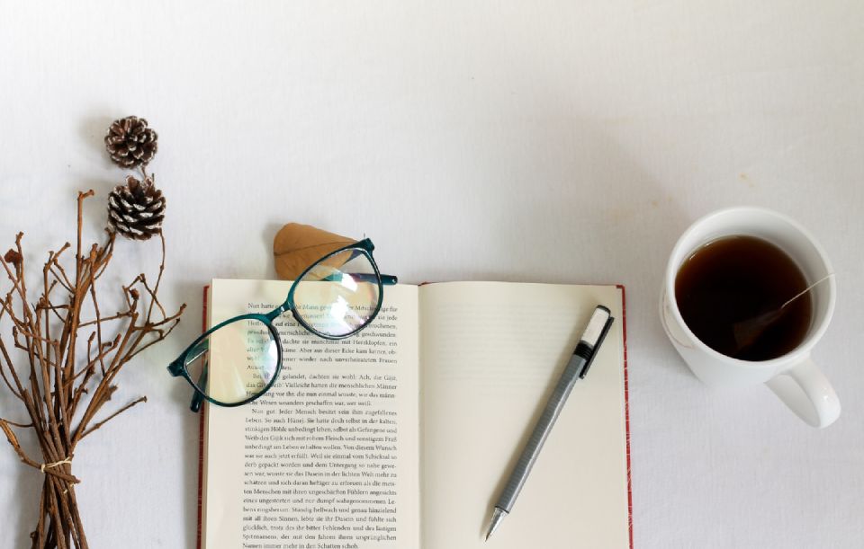 um livro aberto, uma caneta, uma xicara com café, sobre o livro está a caneta e um óculos de grau e ao lado um óculos de sol